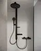 Schwarzes Duschsystem mit Kopfbrause und Handbrause, zwei Ablagen, montiert an Wand mit hellem, strukturiertem Untergrund. Hersteller: Ideal Standard.